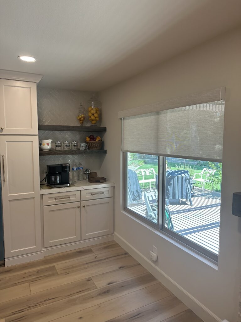 Photo shows a kitchen space with a large window that has a cordless roller shade in a grey/white color that is half up with a custom valance. 